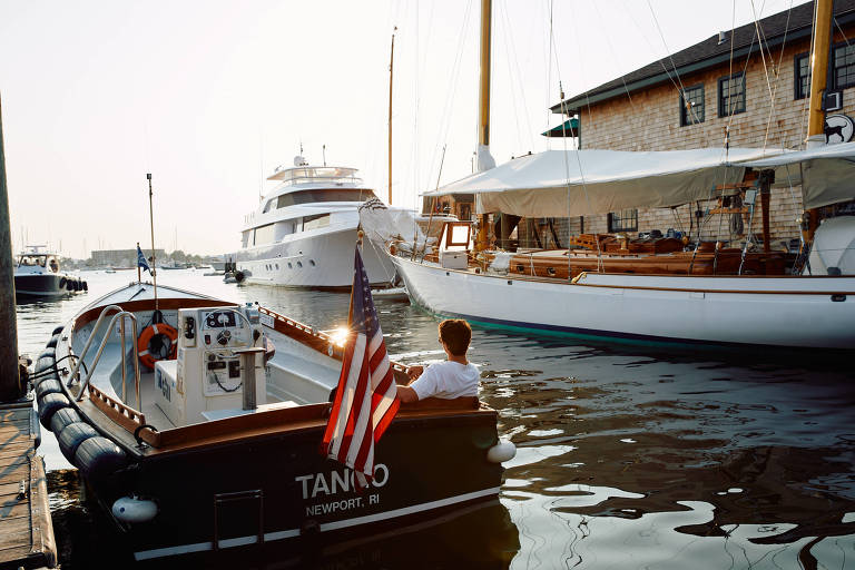 A imagem mostra um pequeno barco de motor ancorado em um porto， com uma pessoa sentada na parte de trás. O barco exibe uma bandeira americana e tem o nome 039;TANTO039; e a localização 039;NEWPORT， RI039; visíveis. Ao fundo， há um iate maior e um barco à vela， além de um edifício de madeira. A luz do sol ilumina a cena， refletindo na água.