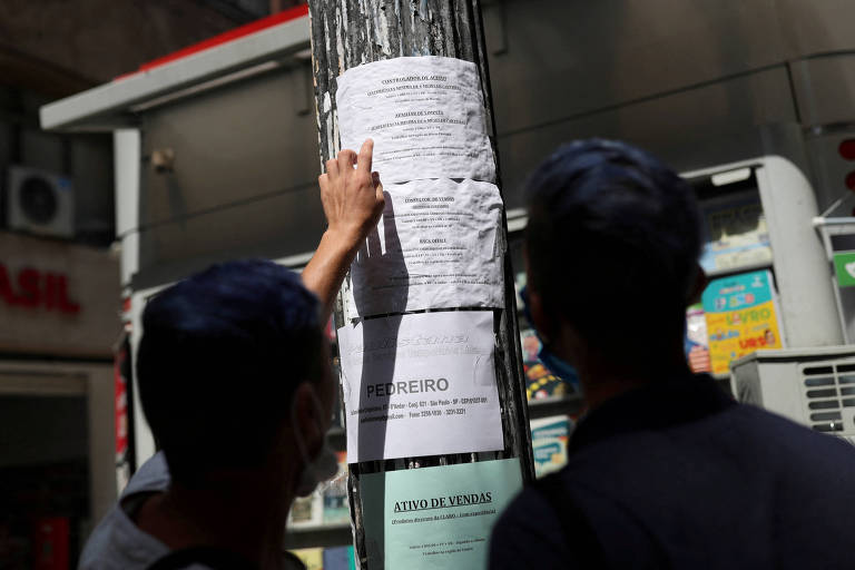 A imagem mostra duas pessoas em frente a um poste de madeira， onde um cartaz branco está colado. Uma das pessoas está com a mão levantada， tocando o cartaz. Ao fundo， há uma loja com letreiro visível e um ambiente urbano. A luz do sol ilumina a cena.