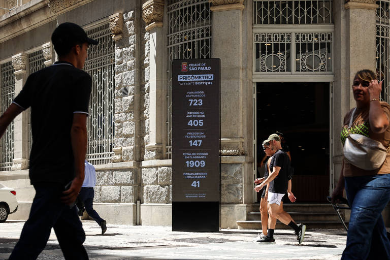 A imagem mostra um painel informativo em uma calçada em São Paulo， com pessoas caminhando ao fundo. O painel exibe dados numéricos， incluindo 723， 405， 147， 1909 e 41. O ambiente é urbano， com uma arquitetura de edifícios antigos ao fundo.