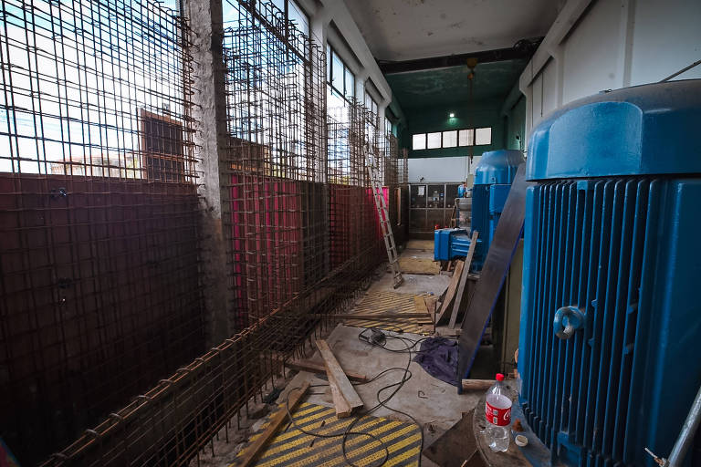 A imagem mostra o interior de um espaço industrial com paredes de concreto e janelas de vidro. À esquerda， há uma grade de metal e， à direita， duas máquinas azuis grandes. O chão é coberto por tábuas de madeira e há uma garrafa de água e fios elétricos visíveis. A iluminação é fraca， com uma luz verde no teto.