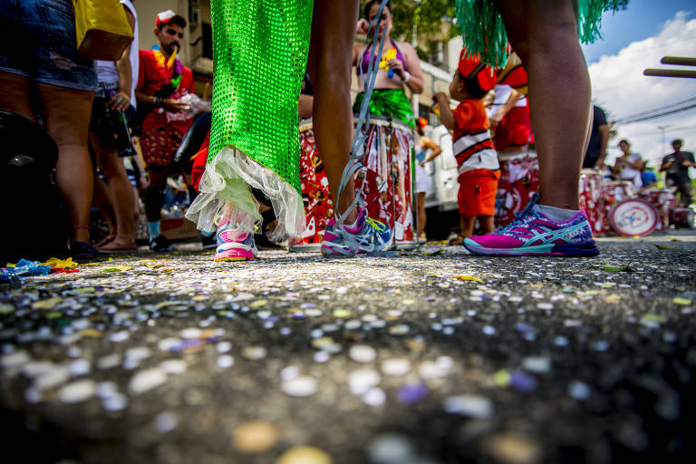 A imagem mostra os peacute;s de vaacute;rias pessoas em um ambiente de carnaval. No primeiro plano， haacute; um par de tecirc;nis coloridos， um verde e um rosa， cercados por confetes espalhados pelo chatilde;o. Ao fundo， eacute; possiacute;vel ver pessoas vestidas com fantasias coloridas e alegres， algumas segurando instrumentos musicais. O cenaacute;rio eacute; festivo e vibrante， tiacute;pico de uma celebraccedil;atilde;o de carnaval.