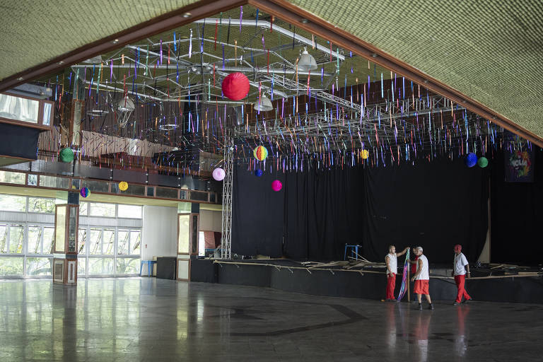 Uma grande sala de eventos com um palco ao fundo. O teto é decorado com fitas coloridas e bolas de papel em várias cores. No chão, três pessoas vestindo roupas brancas e calças vermelhas caminham em direção ao fundo da sala. As paredes têm janelas grandes que permitem a entrada de luz natural.