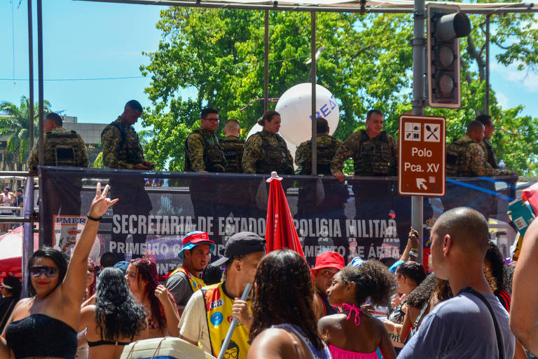 A imagem mostra um evento ao ar livre com uma multidão de pessoas. No fundo, há uma faixa preta com o texto 039;SECRETARIA DE ESTADO DE POLÍCIA MILITAR039; e outros detalhes. Acima da faixa, estão vários homens em uniformes militares, observando a multidão. As pessoas na frente estão vestidas de forma colorida, algumas levantando as mãos e outras dançando. O ambiente é ensolarado, com árvores ao fundo.