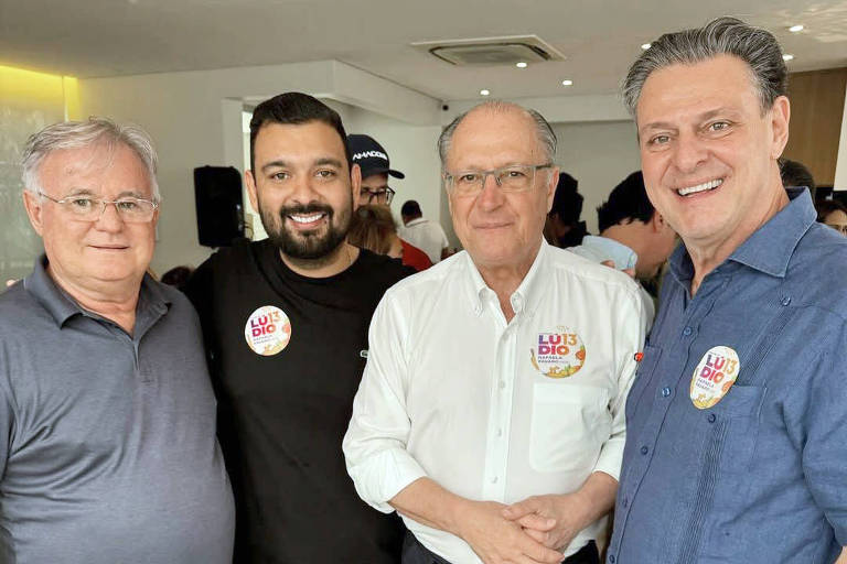 Quatro homens estão posando para a foto em um ambiente interno. Eles estão sorrindo e usando camisetas casuais. Todos estão usando adesivos em seus peitos com a frase 039;BEM VINDO039;. O homem à esquerda está vestindo uma camisa cinza, o segundo está com uma camiseta preta, o terceiro está usando uma camisa branca e o último está com uma camisa azul. O ambiente parece ser um evento social.