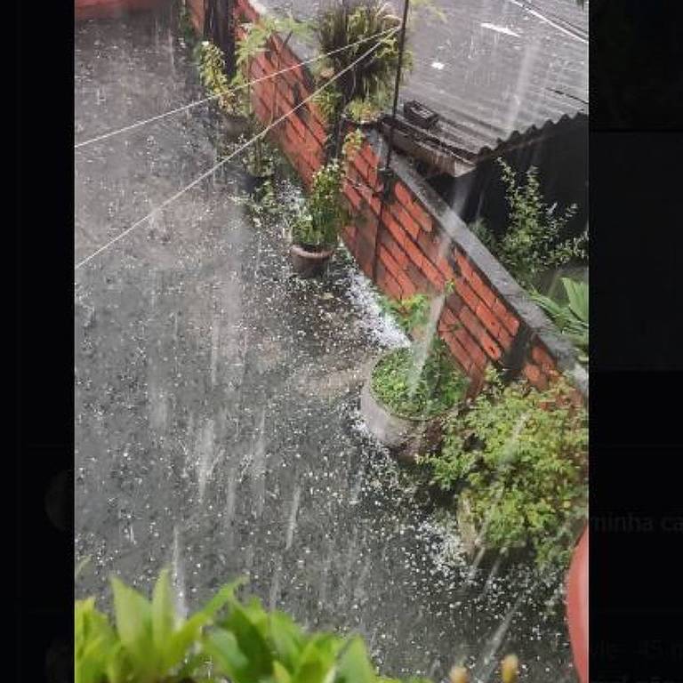 A imagem mostra um pátio coberto por granizo， com plantas em vasos e um muro de tijolos ao fundo. A chuva é intensa， e a água se acumula no chão. É possível ver uma parte de uma casa e uma moto estacionada ao fundo.