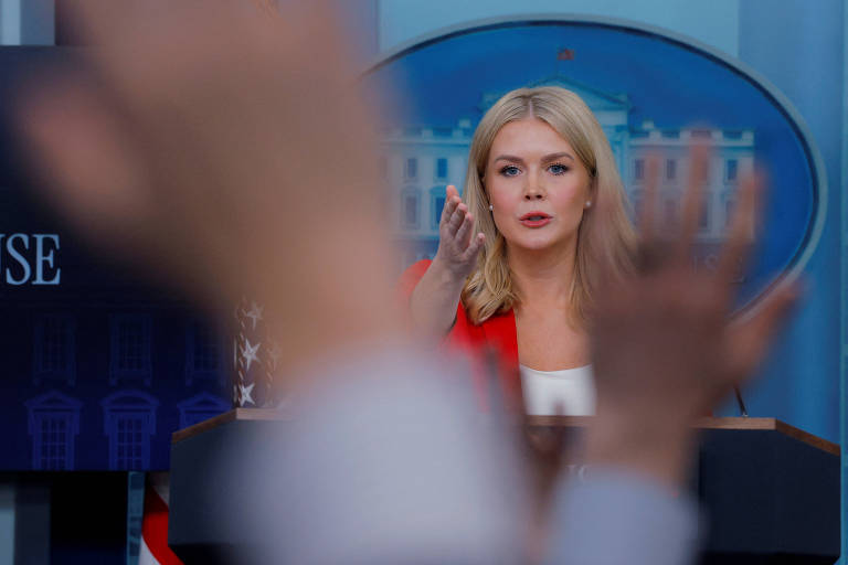 A imagem mostra uma mulher com cabelo loiro e liso， vestindo uma blusa branca e um blazer vermelho， falando em um púlpito durante uma coletiva de imprensa. Ao fundo， é possível ver uma bandeira dos Estados Unidos e uma imagem do edifício da Casa Branca. Mãos de pessoas na audiência estão levantadas， sugerindo que estão fazendo perguntas.