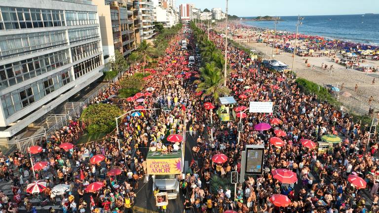 A imagem mostra uma grande multidão de pessoas participando de um evento festivo na praia. Há muitos guarda-sóis vermelhos e uma carreta de som com a frase 'SIMPATIA: É QUASE AMOR' visível. O cenário inclui prédios ao fundo e a praia com várias pessoas e guarda-sóis coloridos. O clima é de celebração e agitação.