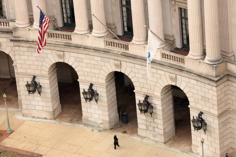 A imagem mostra a fachada de um edifício governamental com colunas imponentes. Há bandeiras dos Estados Unidos e uma bandeira branca visíveis. Abaixo das colunas， há arcos com luminárias penduradas. Uma pessoa está caminhando na calçada em frente ao edifício.