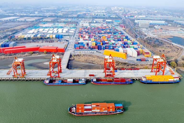 Imagem aérea de um porto com vários navios cargueiros atracados. Há guindastes vermelhos e amarelos operando， e uma grande quantidade de contêineres coloridos empilhados em um terminal próximo. O ambiente é urbano， com áreas industriais visíveis ao fundo.