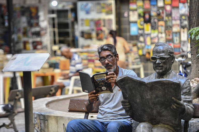 Um homem sentado em um banco de praça lê um livro， enquanto ao seu lado há uma escultura do poeta Mauro Mota sentado， também lendo. Ao fundo， há uma parede coberta de livros e outras pessoas em mesas， criando um ambiente de leitura e convivência.