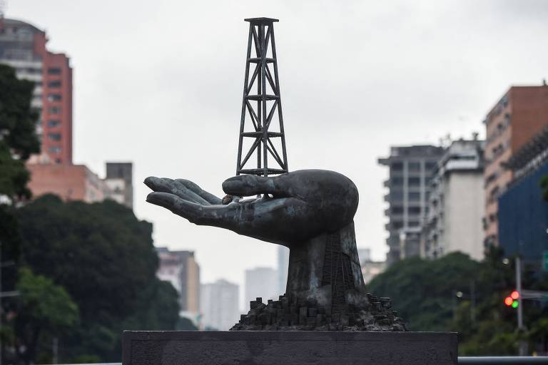 A imagem mostra uma escultura de uma mão humana em bronze， que sustenta uma torre de petróleo. A escultura está posicionada em um espaço urbano， com prédios ao fundo e uma atmosfera nublada. A mão é grande e detalhada， enquanto a torre é mais esguia e se destaca em relação à mão.