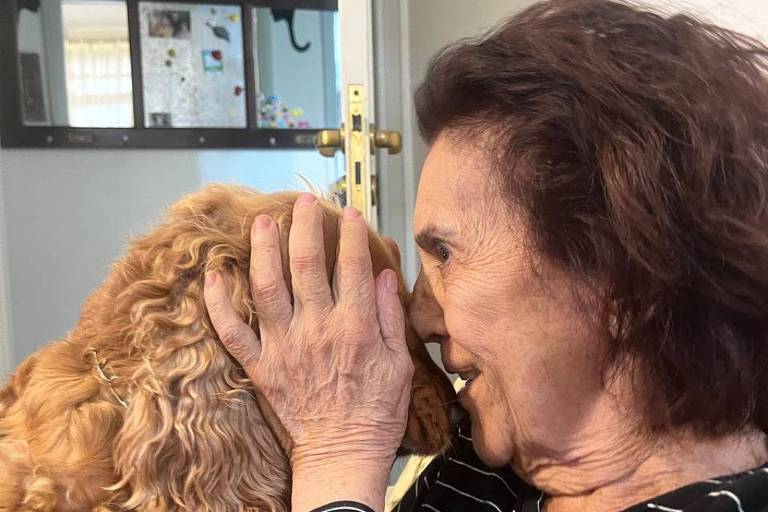 A imagem mostra uma mulher sorridente com cabelo castanho e ondulado， usando uma blusa listrada em preto e branco， segurando um cachorro de pelagem dourada perto de seu rosto. Eles estão em um ambiente interno， com uma porta ao fundo e uma parede decorada. A mulher parece estar brincando ou interagindo carinhosamente com o cão.