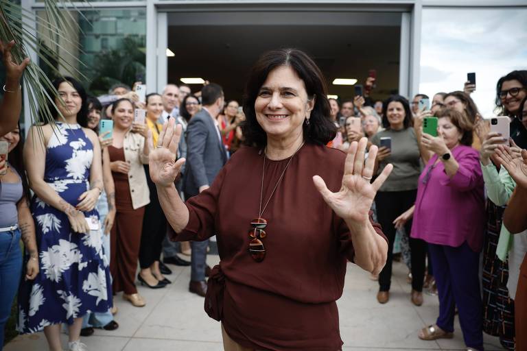 Uma mulher sorridente está em primeiro plano， levantando as mãos em um gesto de saudação. Ela usa uma blusa marrom e um colar. Atrás dela， há uma multidão de pessoas， algumas aplaudindo e outras segurando celulares para registrar o momento. O ambiente parece ser ao ar livre， com uma entrada de prédio ao fundo.