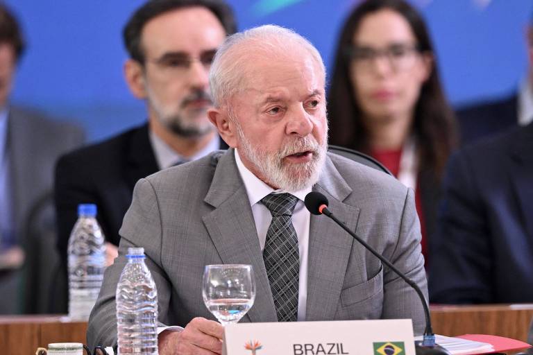 Um homem de cabelo grisalho e barba, vestido com um terno cinza e gravata, fala em um microfone durante uma reunião. À sua frente, há um copo de água e uma garrafa de água. Ao fundo, outras pessoas estão sentadas, algumas olhando para ele. Uma bandeira do Brasil é visível na mesa.