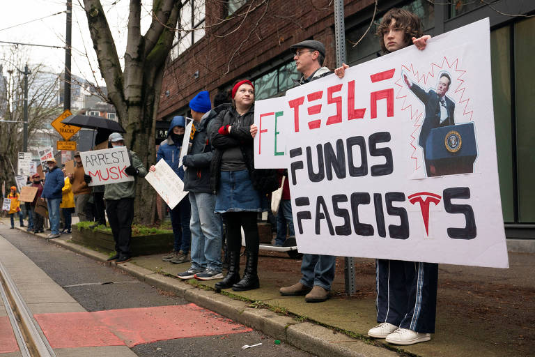 A imagem mostra um grupo de pessoas protestando na calçada. Algumas pessoas seguram cartazes， incluindo um que diz 039;Fora Tesla039; e 039;Tesla financia Fascistas039;， com uma ilustração de uma figura apontando. Os manifestantes estão vestidos com roupas de inverno， e há uma variedade de chapéus e cachecóis. Ao fundo， há um edifício e sinais de trânsito.