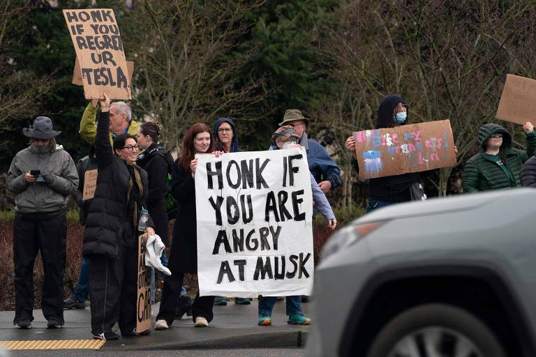 Um grupo de pessoas participa de um protesto em uma rua, segurando cartazes. Um dos cartazes principais diz 'HONK IF YOU ARE ANGRY AT MUSK'. Outros cartazes têm mensagens variadas. Algumas pessoas estão usando máscaras. Ao fundo, há árvores e um carro passando.