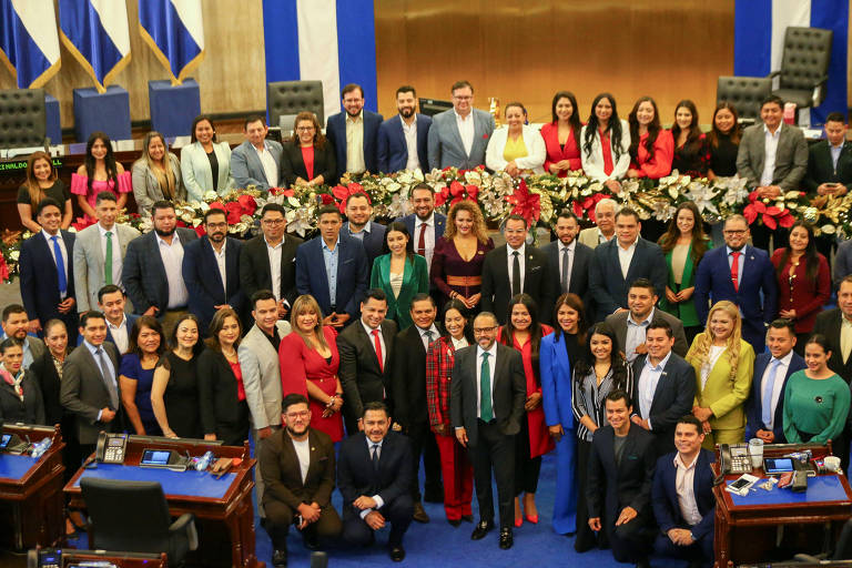 Uma grande foto em grupo de pessoas em um ambiente legislativo. O fundo apresenta bandeiras e uma decoração festiva com flores. As pessoas estão vestidas de maneira formal e variada， com algumas usando roupas coloridas. O grupo é composto por homens e mulheres， e todos estão sorrindo para a câmera.