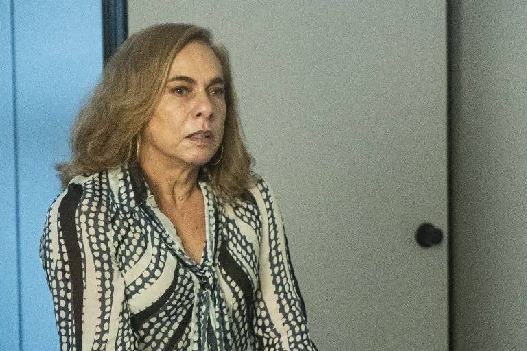 Uma mulher está em pé ao lado de uma cadeira em um ambiente de escritório. Ela tem cabelo castanho claro e usa uma blusa com padrão de listras em preto e branco. A expressão dela parece séria ou preocupada. O fundo é de um tom neutro, com uma porta e uma parede visíveis.