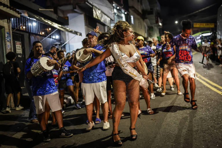 A imagem mostra um grupo de pessoas participando de um desfile de carnaval à noite. No centro， uma mulher com cabelo cacheado e vestido curto dança， segurando uma faixa. Ao seu redor， homens e mulheres tocam instrumentos de percussão e dançam， todos vestindo camisetas coloridas. O ambiente é urbano， com luzes e edifícios ao fundo.