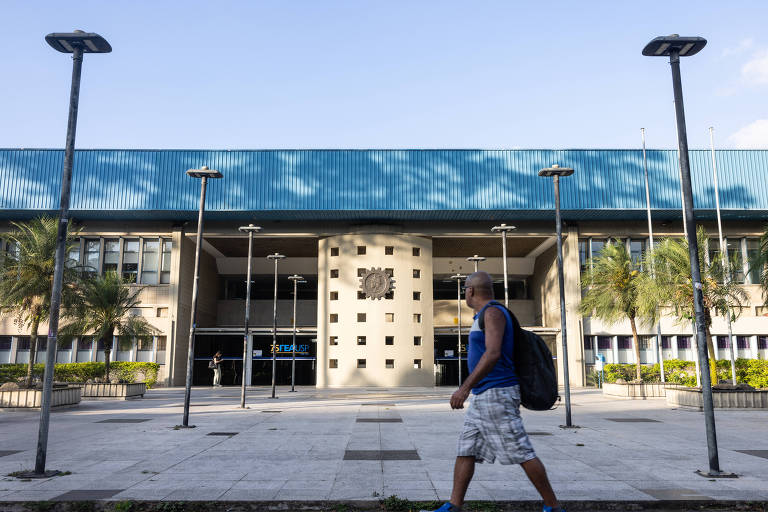 A imagem mostra a fachada de um edifício público com um telhado azul. Na frente， há um homem caminhando， vestindo uma camiseta escura e shorts claros. Há postes de luz ao longo da calçada. O céu está claro e há algumas sombras projetadas no chão.