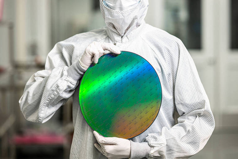 Um profissional vestido com um traje de proteção， incluindo máscara e luvas， está segurando um wafer de silício. O wafer tem uma superfície refletiva com cores iridescentes， como verde e azul. Ao fundo， há um ambiente de laboratório com portas e janelas.