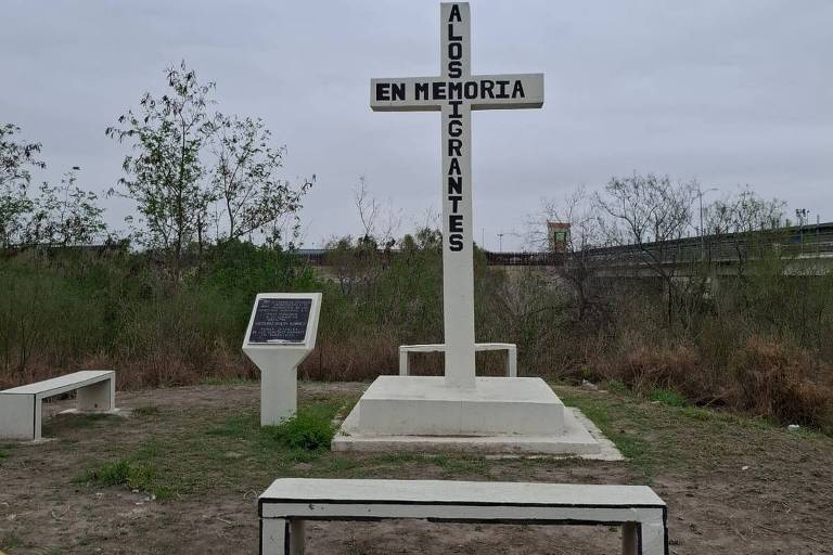 A imagem mostra uma cruz branca em um espaço aberto， cercada por vegetação baixa. A cruz possui a inscrição 039;A LOS MIGRANTES EN MEMORIA039; em letras pretas. À esquerda da cruz， há um painel informativo em uma estrutura branca. No chão， há bancos brancos dispostos em frente à cruz. O céu está nublado， sugerindo um clima sombrio.