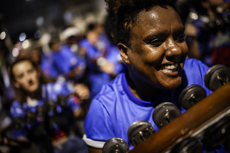 A imagem mostra um grupo de pessoas em um evento festivo, com foco em uma mulher sorridente que toca um instrumento musical. Ela está usando uma camiseta azul e parece estar se divertindo. Ao fundo, outras pessoas também estão vestidas de azul e participando da celebração.