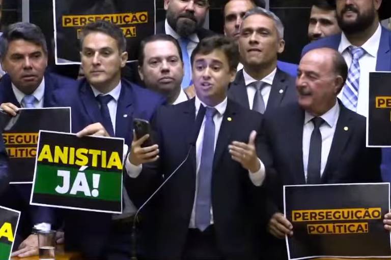 A imagem mostra um grupo de homens em um plenário， muitos deles usando terno e gravata. No centro， o deputado Nikolas Ferreira está falando e segurando um cartaz que diz 039;ANISTIA JÁ!039;. Outros membros ao fundo seguram cartazes com a frase 039;PERSEGUIÇÃO POLÍTICA039;. A cena parece ser de uma sessão deliberativa na Câmara dos Deputados.