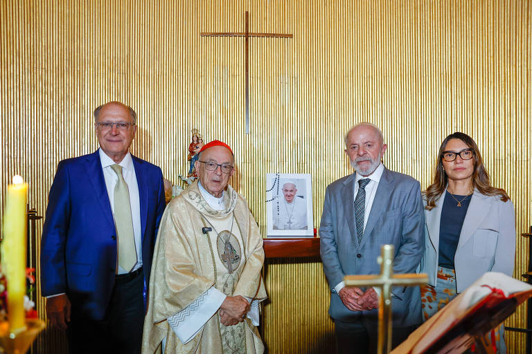 A imagem mostra quatro pessoas em uma cerimônia religiosa. No centro， um cardeal vestido com vestes litúrgicas. À sua esquerda， um homem de terno azul e gravata clara. À direita， um homem de terno cinza e gravata clara. Uma mulher com óculos e blazer claro está ao lado do homem de terno cinza. Ao fundo， uma parede dourada com uma cruz e uma imagem de um papa em um suporte.