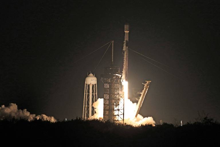 A imagem mostra um foguete sendo lançado à noite， com chamas e fumaça saindo da base. O foguete está em pé sobre uma plataforma de lançamento， com estruturas metálicas ao redor. O céu está escuro， destacando a luz intensa do lançamento.