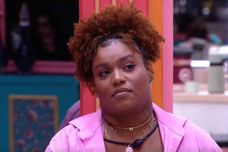 A imagem mostra uma mulher com cabelo cacheado e volumoso, usando uma blusa rosa. Ela está com uma expressão séria e parece estar em um ambiente interno, com paredes coloridas ao fundo. Há objetos e pessoas desfocados ao fundo, sugerindo um ambiente social.