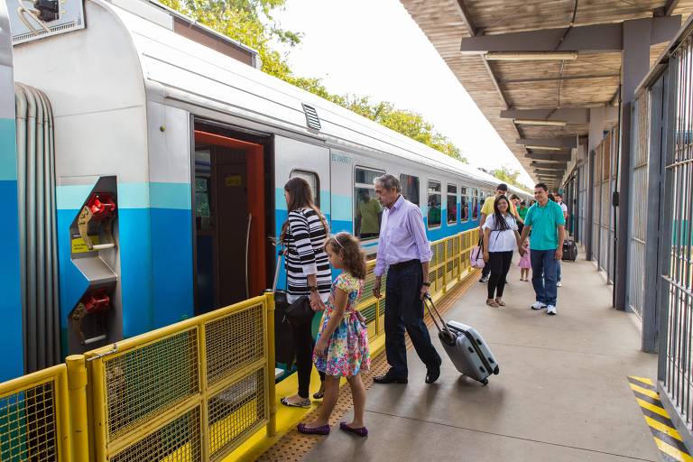 A imagem mostra um grupo de pessoas embarcando em um trem em uma estação. Há uma mulher com uma mala， uma criança com um vestido colorido， um homem de camisa branca e calças escuras， e outras pessoas ao fundo. O trem é de cor azul e branca， e a plataforma é ampla， com cercas de segurança. O ambiente é iluminado e parece ser um dia ensolarado.