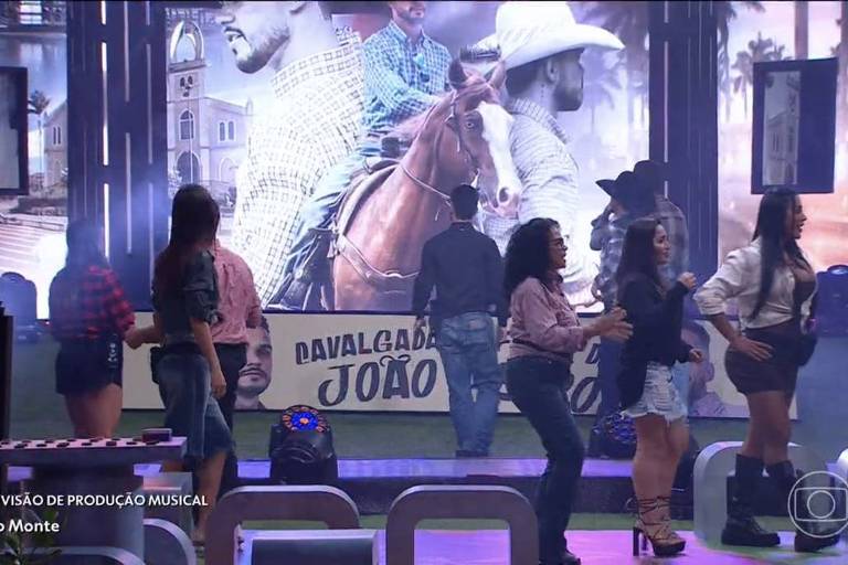 A imagem mostra um palco de apresentação musical com várias dançarinas em trajes típicos, dançando em grupo. Ao fundo, há um grande banner com a imagem de um homem montado em um cavalo, e o texto 'NIVALCADÃO JOÃO'. A iluminação é colorida e o ambiente parece festivo.