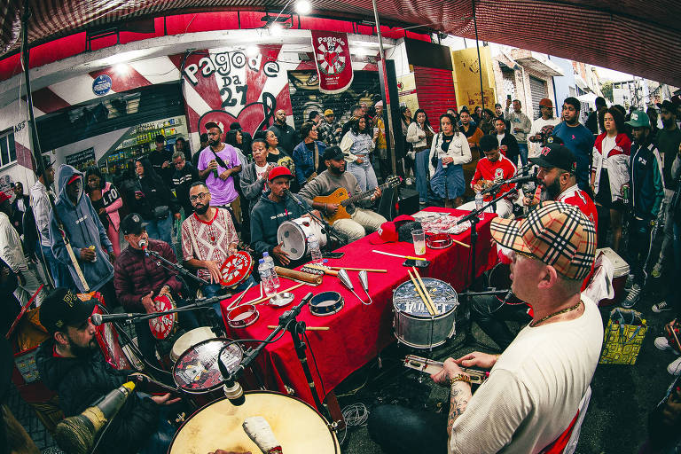 A imagem mostra um grupo de músicos tocando instrumentos de percussão em um espaço público coberto. Há uma mesa vermelha à frente， com vários instrumentos e objetos. O público， composto por várias pessoas， observa atentamente a apresentação. O ambiente é festivo， com uma decoração colorida e uma tenda sobre os músicos.