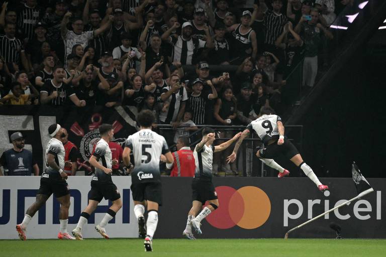 Um grupo de jogadores de futebol celebra um gol em um estádio lotado. Um jogador， identificado pelo número 9， está pulando em direção a um canto do campo， enquanto outros jogadores se aproximam para comemorar. Ao fundo， a torcida está animada， vestindo camisas listradas em preto e branco. Há um banner publicitário visível na parte inferior da imagem.