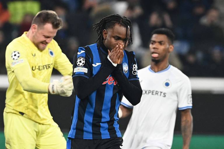 O atacante Ademola Lookman， do Atalanta， leva as mãos ao rosto， cobrindo com elas boca e nariz， ao lamentar pênalti desperdiçado contra o Brugge na Champions League; atrás dele， o goleiro Mignolet comemora