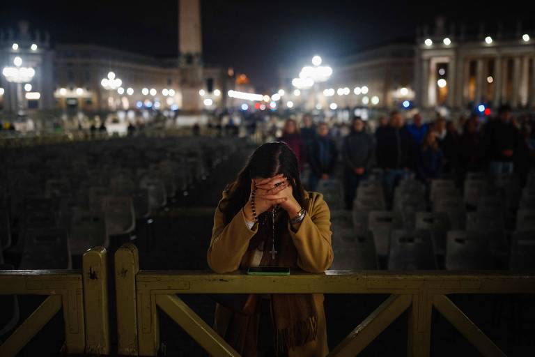 A imagem mostra uma mulher em um momento de reflexão ou tristeza， com as mãos cobrindo o rosto. Ela está em frente a uma grade， em uma praça iluminada à noite， onde há várias cadeiras vazias. Ao fundo， é possível ver um obelisco e outras pessoas em pé， também em silêncio. A iluminação da praça cria um ambiente solene.