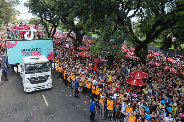 A imagem mostra uma grande multidão reunida em uma rua durante um evento de carnaval， com um caminhão de som na frente. O caminhão é decorado com a marca do TikTok e tem pessoas em cima dele. A multidão é composta por pessoas usando camisetas laranjas e segurando guarda-chuvas vermelhos. Árvores estão visíveis ao longo da rua.