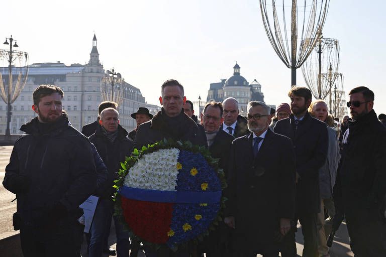 Um grupo de pessoas está reunido em um local ao ar livre, possivelmente em uma cerimônia. No centro, um homem segura uma coroa de flores que tem as cores da bandeira da Rússia e da União Europeia. Ao fundo, há edifícios e estruturas urbanas. As pessoas estão vestidas de forma variada, algumas em trajes formais. O clima parece ensolarado.