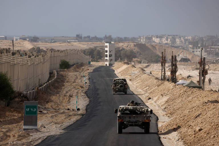 A imagem mostra uma estrada pavimentada em uma área desértica， com veículos militares se deslocando. À esquerda， há uma cerca alta e uma estrutura de segurança. Ao fundo， é possível ver uma construção e uma paisagem árida， com algumas árvores esparsas e postes de energia.