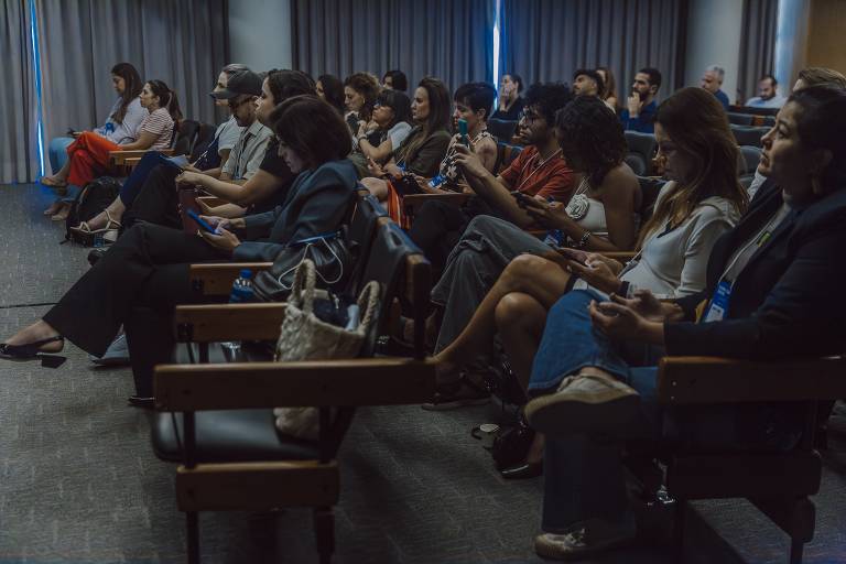 A imagem mostra um público sentado em um auditório. As pessoas estão atentas, algumas usando dispositivos móveis. O ambiente é bem iluminado, com cortinas cinzas ao fundo. Há uma diversidade de estilos de roupas entre os participantes, que incluem tanto roupas formais quanto casuais.