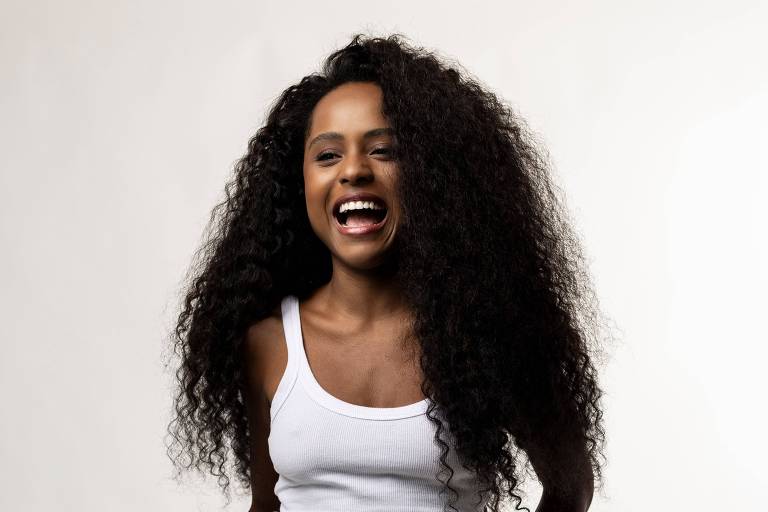 A imagem mostra uma mulher com cabelo longo e cacheado, usando uma camiseta branca sem mangas e calças largas. Ela está sorrindo e posando de forma descontraída, com uma das mãos na cintura e a outra levemente levantada. O fundo é claro e neutro