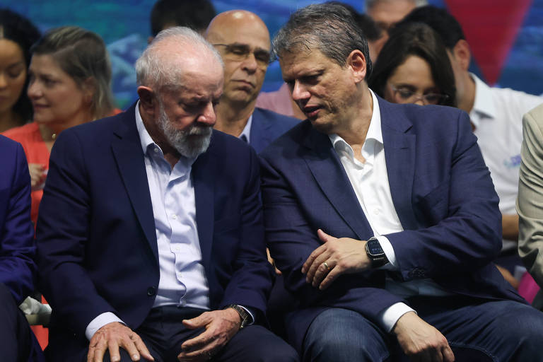 A imagem mostra um grupo de pessoas sentadas em um evento. Em primeiro plano， dois homens estão conversando. O homem à esquerda tem cabelo grisalho e usa um terno escuro， enquanto o homem à direita tem cabelo curto e usa um terno claro. Ao fundo， outras pessoas estão visíveis， algumas olhando para frente e outras interagindo entre si.