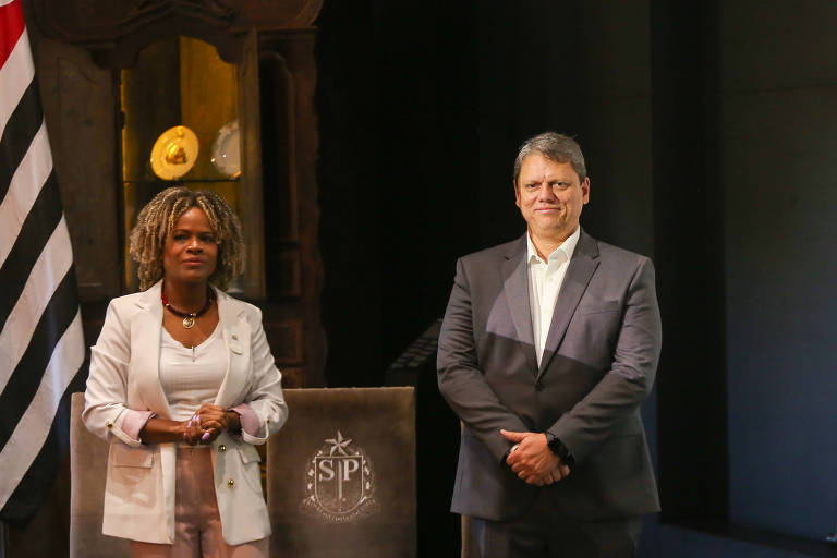 A imagem mostra duas pessoas em um evento oficial. À esquerda， Helena Reis， uma mulher com cabelo cacheado e vestido claro， usando um blazer branco e acessórios. À direita， Tarcísio de Freitas， um homem com cabelo curto e grisalho， vestido com um terno escuro e camisa clara. Ao fundo， há uma bandeira do estado de São Paulo e um painel decorativo com o brasão do estado.