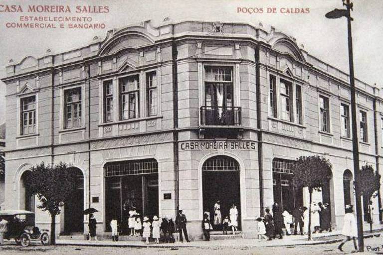 A imagem em preto e branco mostra a fachada da Casa Moreira Salles, um estabelecimento comercial e bancário localizado em Poços de Caldas. O edifício é de estilo arquitetônico clássico, com janelas grandes e detalhes ornamentais. Na calçada, há várias pessoas, algumas vestindo roupas de época, e um carro antigo estacionado em frente ao prédio. O nome 'CASA MOREIRA SALLES' está visível na fachada.