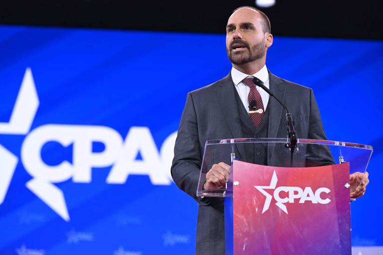 Um homem em um terno escuro está falando em um púlpito transparente com o logotipo do CPAC em destaque. O fundo é azul com o logotipo do CPAC em branco. O homem tem cabelo curto e barba bem aparada， e parece estar se dirigindo a uma audiência.