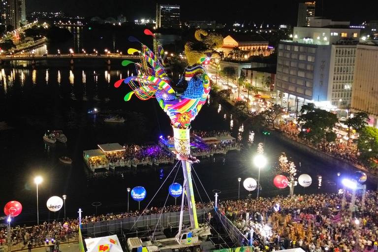 A imagem mostra a escultura gigante do Galo da Madrugada no Recife， com um rio ao centro e muitos edifícios iluminados ao fundo. Há uma grande estrutura colorida no centro da cena， com várias pessoas reunidas ao redor. Balões coloridos e luzes decorativas estão presentes， criando um ambiente festivo. O céu está escuro， indicando que é noite.