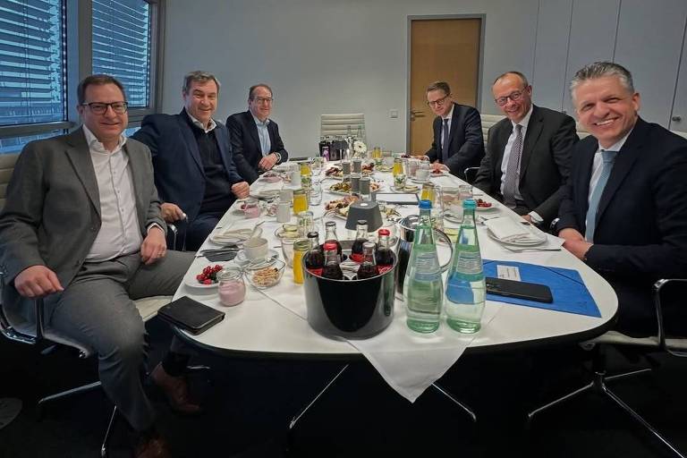 A imagem mostra um grupo de seis homens sentados ao redor de uma mesa de conferência. A mesa está coberta com toalhas brancas e contém garrafas de água， copos， e algumas bebidas em latas. Ao fundo， há janelas com persianas e uma porta. Os homens estão sorrindo e parecem estar em um ambiente de negócios.