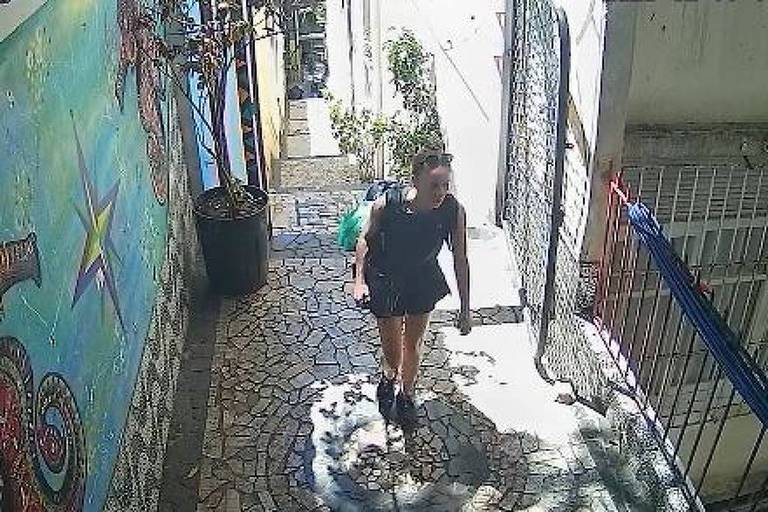 A imagem mostra uma mulher caminhando por um beco estreito， com paredes decoradas por um mural colorido. O chão é coberto por pedras， e há uma planta em um vaso à esquerda. A mulher está vestindo uma blusa preta e shorts， carregando uma mochila nas costas. A luz do sol ilumina a cena， criando sombras no chão.