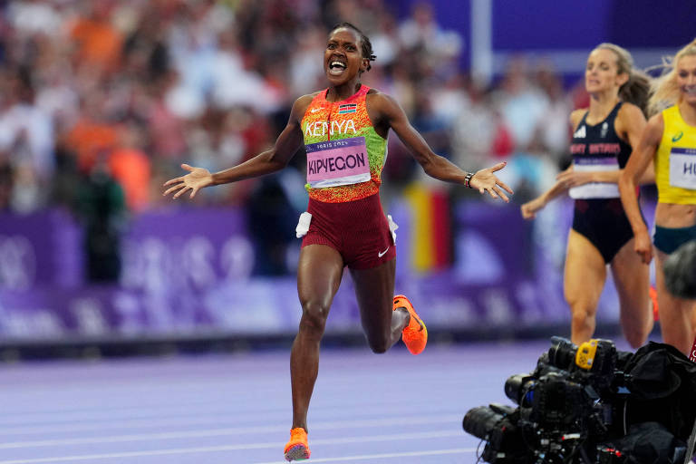 Uma atleta está correndo na pista de atletismo， com os braços abertos em celebração. Ela usa um uniforme vermelho e amarelo com a palavra 039;KENYA039; em destaque. Ao fundo， outras competidoras estão correndo， e uma multidão é visível nas arquibancadas.